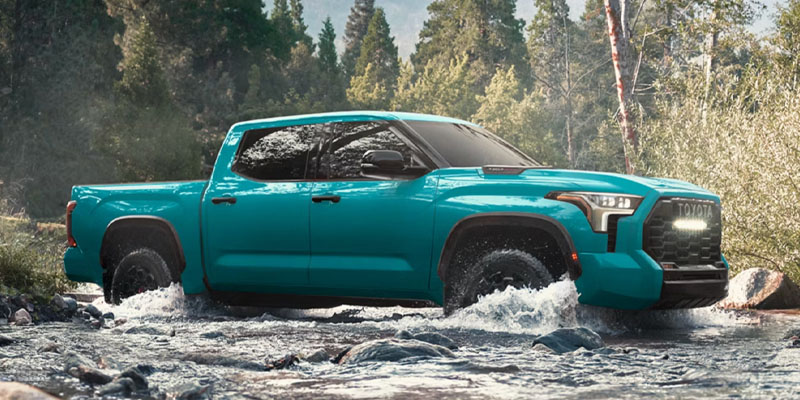 2026 Toyota Tundra pickup truck in teal driving through a shallow river, splashing water as it navigates a rocky forest setting in Frederick, Colorado.