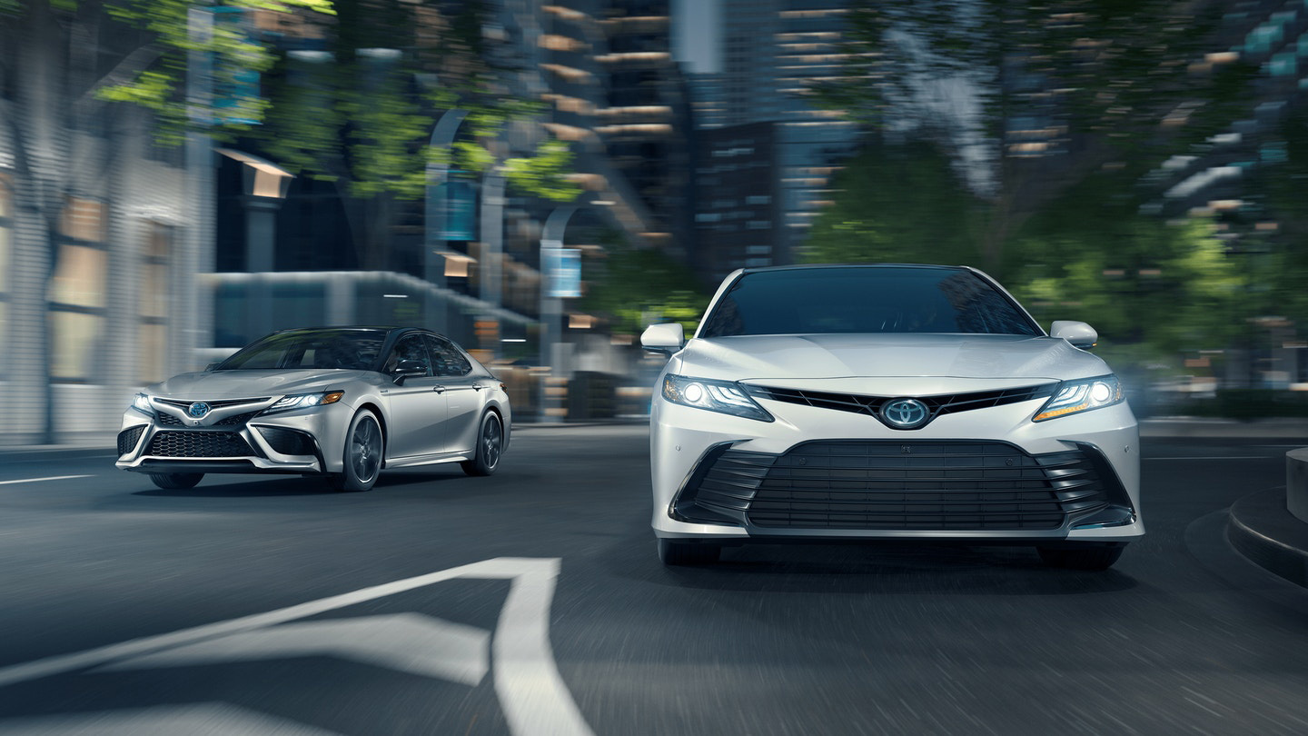 2026 Toyota sedans driving through a modern city street, shown in front and side views, featuring sleek headlights, bold front grilles, and motion blur from surrounding buildings and trees.