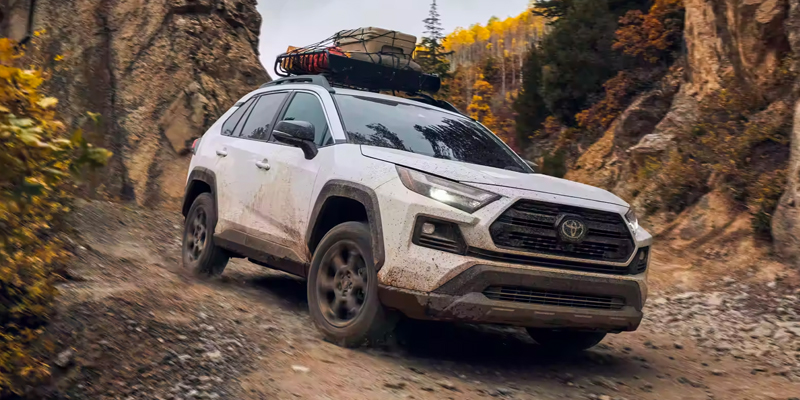 A Toyota RAV4, covered in dirt and mud, driving through a rugged mountain trail with a roof rack loaded with gear, showcasing its off-road capability.