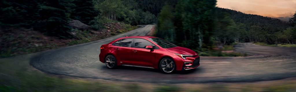 A red Toyota Sedan driving on a winding road surrounded by trees and mountains.
