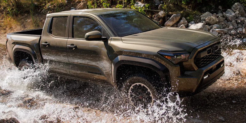 A Toyota Tacoma truck driving through a shallow stream, splashing water. The vehicle is in an outdoor setting with rocks and greenery around.